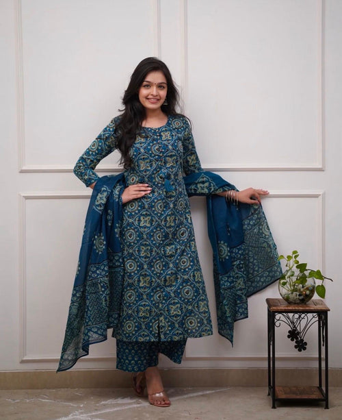 Woman wearing a blue patterned traditional outfit standing against a white paneled wall.